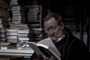 Older man sat reading a book among a pile of books